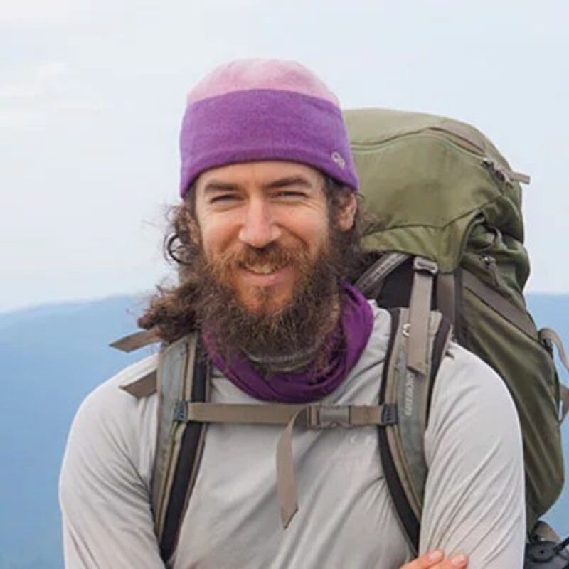 The image shows a man with a long beard and a purple and pink beanie. He is wearing a light-colored long-sleeved shirt and a purple scarf. He has a large backpack on, suggesting he is hiking or backpacking. The background is a blurred landscape, possibly mountains.